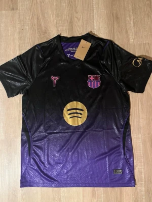 Barcelona borttröja Yamal Lila/Svart 26/27 - Snygg FC Barcelona bortatröja i svart och lila med guldiga detaljer och tryck. Inför säsongen 26/27. Tröjan har Lamine Yamal och nummer 10 på ryggen, samt Barca Foundation-logga. Tillverkad i ett lätt och ventilerande material, perfekt för fotboll eller träning. HELT NY!
