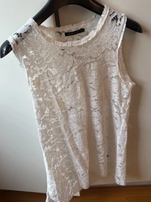 Vit spetsärmlös topp från Mirelle - Säljer en vit ärmlös topp från Mirelle i vacker spets med blommigt mönster. Perfekt för dig som gillar romantisk stil och vill ha något lätt och luftigt till sommaren. Toppen har rund halsringning och är transparent.