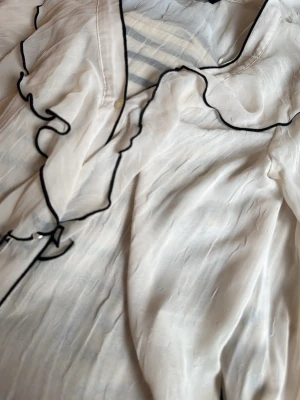 Vit blus med volang och svarta kanter - Snygg vit blus i tunn, lätt material med stora volanger längs kragen och knäppning framtill. Svarta kontrastsömmar ger en cool touch. Perfekt för dig som gillar romantisk och trendig stil. Från Zara