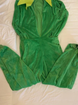 Grön Kermit-onesie med huva - Rolig och mjuk Kermit-maskeraddräkt i grön plysch med dragkedja framtill. Dräkten har huva med Kermits ansikte, stora ögon samt gula detaljer runt halsen. Perfekt för cosplay eller maskerad!