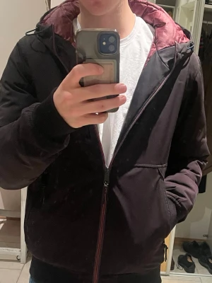 Dunjacka från Scotch & Soda - Säljer en svart pufferjacka från Scotch & Soda i storlek S. Jackan har vinrött quiltat foder, huva och två snedställda fickor med dragkedja. Perfekt för kalla dagar och har en clean look med diskreta detaljer.