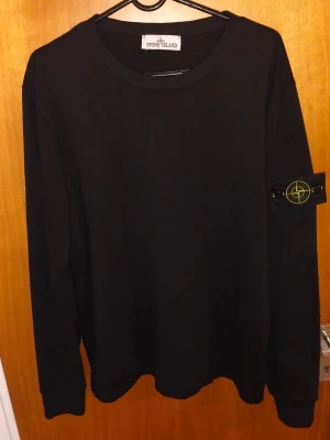 Svart långärmad topp från Stone Island - Svart långärmad topp från Stone Island med klassisk rund hals och ikonisk patch på ärmen. Tillverkad i mjuk bomull för en clean och stilren look. Perfekt för dig som gillar minimalistisk stil och streetwear-vibe.