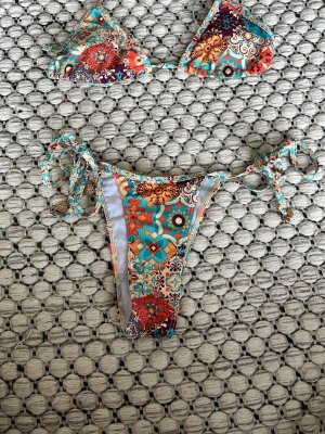 Färgglad blommig bikini med knyt - Säljer en färgglad bikini med blommigt mönster i turkos, orange, röd och beige. Överdelen har trekantsform och underdelen har knyt i sidorna. Perfekt för sommaren och strandhäng! Helt nytt skick. 1 del = 70 kr, 2 delar = 120 kr