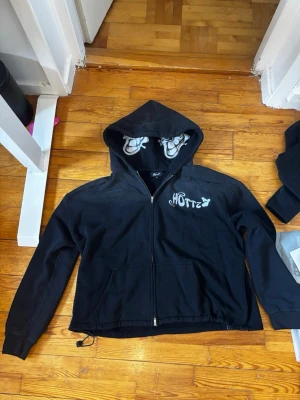 Svart zip hoodie med tryck - Säljer en svart hoodie med dragkedja och huva. Tryck med texten 'HOTTZ' på bröstet och blommönster i luvan. Justerbar dragsko i nederkanten och två fickor framtill. Perfekt för en chill och avslappnad stil. sitter super stort passar mest för bredare personer