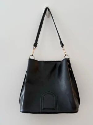 Bucket bag  - Svart handväska i bucket-modell med guldfärgade metalldetaljer och avtagbart axelband. Väskan har en enkel, slät design med dekorativ söm framtill och två nitar på sidan. Perfekt för dig som gillar minimalistisk och modern stil.