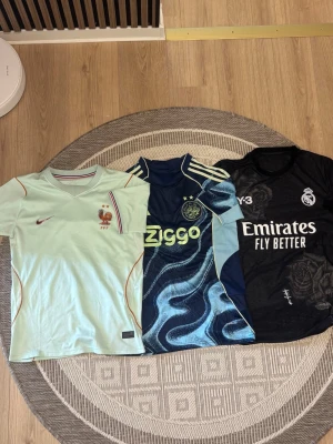 Tre fotbollströjor Nike, Adidas, Y-3 - Säljer tre snygga fotbollströjor: en ljusblå Nike-tröja med Frankrikes landslagsmärke, en blå Adidas Ajax-tröja med Ziggo-logga och vågmönster samt en svart Real Madrid-tröja från Y-3 med vita detaljer och tryck. Alla har kort ärm och sportig passform.