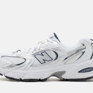 Vita New Balance 530 sneakers - Klassiska vita New Balance 530 sneakers med meshpaneler och detaljer i silver och mörkblått. Skorna har en chunky sula med ABZORB-dämpning och tydlig logga på sidan. Perfekta för en sportig och trendig look.