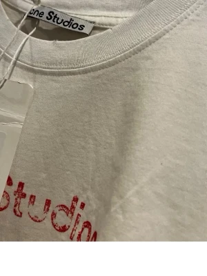 Vit Acne Studios t-shirt i bomull - Säljer en vit t-shirt från Acne Studios i 100% bomull. T-shirten har rund halsringning och tryckt röd text på bröstet. Enkel och clean design som passar perfekt till jeans eller shorts.