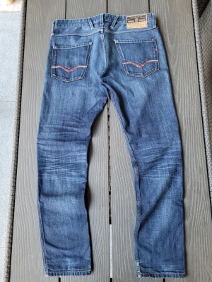 Blå replay anbass - Säljer ett par replay anbass med snygga slitningar och kontrastsömmar på bakfickorna. Jeansen har klassisk femficksdesign, knappgylf och är tillverkade i ett mjukt denimtyg. Perfekta för en avslappnad och trendig look.