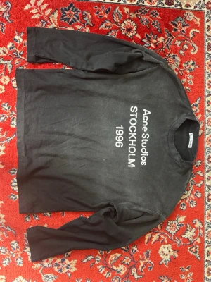 Svart långärmad t-shirt Acne Studios - Svart långärmad t-shirt från Acne Studios med tryck i vitt på framsidan där det står 'Acne Studios STOCKHOLM 1996'. Klassisk rund halsringning och avslappnad passform. Perfekt för en stilren och modern look. Kan inte garantera äktheten 