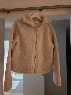 Beige teddyjacka med dragkedja - Mysig beige teddyjacka med hög krage och dragkedja framtill. Jackan har två stora fickor och är gjord i fluffigt teddy-material som håller dig varm. Perfekt för kyliga dagar och ger en avslappnad, trendig look.