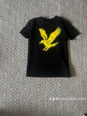 Svart Lyle & Scott t-shirt med gul örn - Snygg svart t-shirt från Lyle & Scott med en stor gul örn på bröstet. Klassisk rund hals och korta ärmar. Perfekt för dig som gillar streetwear och vill sticka ut med en cool print. Tillverkad i mjuk bomull för skön känsla.