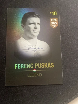 FIFA 365 Ferenc Puskás Legend-kort - Samlarkort från FIFA 365-serien med Ferenc Puskás som legend. Kortet har svart bakgrund med holografiska detaljer och guldtext. Framsidan visar ett porträtt av Puskás och baksidan har en kod för onlineutmaningar. Perfekt för fotbollsfans och samlare.