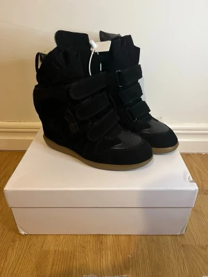 Isabel Marant skor - Svarta sneakers från Isabel Marant med inbyggd kilklack och kardborreband. Skorna har en cool, chunky siluett och är gjorda i mocka och läder med gummisula. Perfekta för dig som vill ha både höjd och streetstyle-vibe.