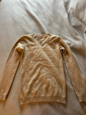 Beige v-ringad cashmeretröja Kujten - Mysig beige stickad tröja från Kujten i mjuk cashmere. Tröjan har v-ringning, långa ärmar och ribbade muddar vid ärmslut och nederkant. Perfekt för lager på lager och enkel att matcha med olika stilar. Helt oanvänd och värde på 2000