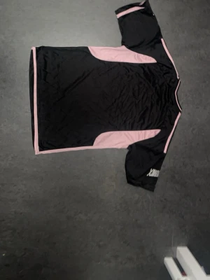 Svart och rosa Inter Miami matchtröja - Snygg svart matchtröja från Inter Miami med rosa detaljer och logga på bröstet. Tröjan har korta ärmar, rund hals och tre rosa ränder på axlarna. Tillverkad i lätt och ventilerande funktionsmaterial, perfekt för fotboll eller träning.