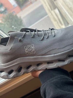 On cloud LOEWE - Snygga grå sneakers med ventilerad mesh-överdel och en chunky, vågig sula med hål för extra komfort. Skon har ett stilrent broderat mönster på sidan och matchande grå skosnören. Perfekt för dig som gillar modern och sportig stil.