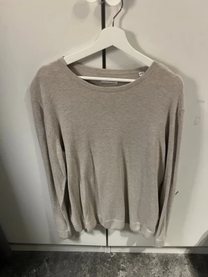 Beige basic tröja med rund hals - En enkel och stilren beige tröja med rund halsringning och långa ärmar. Tröjan har en avslappnad passform och är tillverkad i ett mjukt, tunt material som passar perfekt till lager-på-lager. Perfekt för dig som gillar minimalistisk stil.