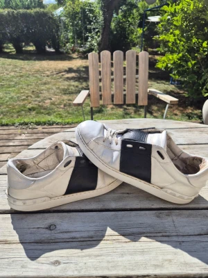 Valentino vita sneakers med svart detalj - Säljer ett par snygga vita sneakers från Valentino med bred svart läderdetalj över sidan och ovandelen. Klassisk låg modell med vita snören och diskret logga på plösen. Perfekta för dig som gillar stilrena och exklusiva skor.