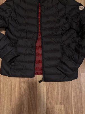 Svart dunjacka från Moncler - Snygg svart dunjacka från Moncler med quiltad design och dragkedja framtill. Jackan har två sidofickor med dragkedja och röd innerfoder med Moncler-logga. Perfekt för kalla vinterdagar och har en klassisk Moncler-patch på ärmen.