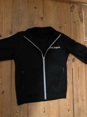 Svart Palm Angels full zip - Säljer en svart hoodie från Palm Angels med vit dragkedja och vit logga på bröstet. Hoodien har vita detaljer längs ärmarna och två fickor framtill. Perfekt för dig som gillar streetwear och vill ha en stilren look.