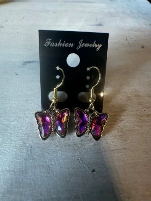 Lila fjärilsörhängen i metall - Säljer ett par snygga örhängen i form av fjärilar med skimrande lila och rosa stenar. Örhängena har guldfärgade krokar och en glansig finish som fångar ljuset. Perfekta för dig som vill ha något unikt och färgglatt till din stil.