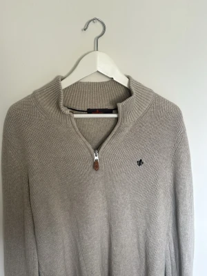 Beige stickad tröja från Morris - Snygg beige stickad tröja från Morris med halv dragkedja och ribbad krage. Tröjan har en diskret logga broderad på bröstet och är tillverkad i mjukt material. Perfekt för lager på lager och enkel att matcha med olika outfits.