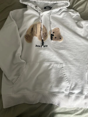 Vit hoodie Palm Angels med björn - Säljer en vit hoodie från Palm Angels med ett broderat björnmotiv på bröstet och svart logga under. Hoodien har stor ficka framtill, ribbade muddar och dragsko i huvan med tryck. Perfekt för dig som gillar streetwear och vill sticka ut.