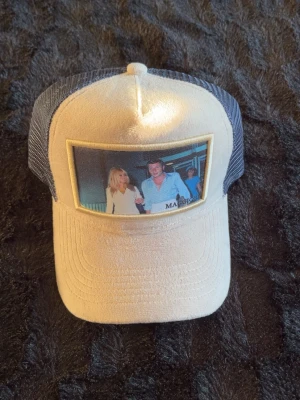 Maggiore Beige och blå keps  - Snygg truckerkeps i beige med blå mesh på sidorna och baksidan. Framtill sitter ett foto med två personer i festmiljö, vilket ger kepsen en unik och retro vibe. Skärmen är böjd och materialet är en mix av tyg och mesh för extra ventilation.