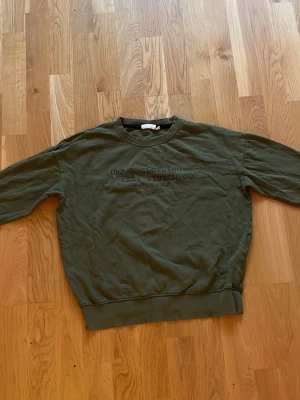 Olivgrön sweatshirt med tryck - Olivgrön sweatshirt med texten 'Urban Clothing Street Vision Co.' tryckt på bröstet. Tröjan har rund halsringning, långa ärmar och ribbade muddar vid ärmslut och nederkant. Perfekt för en avslappnad streetwear-look. Storlek:11-12 år, 146-152. 