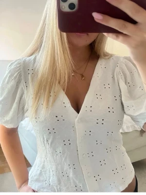 Vit blus - Söt vit blus med v-ringning, puffiga korta ärmar och broderade hål som ger en luftig känsla. Knäpps framtill med små knappar och har en lätt croppad passform. Perfekt för vår och sommar!💓