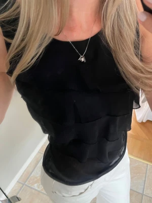 Svart volang topp med knyt - Säljer en svart ärmlös topp med flera lager volanger och knyt på ena axeln. Toppen har en lätt transparent känsla vid magen och sitter snyggt figurnära. 