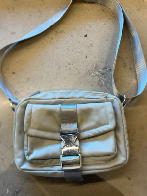 Ljusgrå axelväska med spänne - Snygg ljusgrön axelväska i nylon med justerbar axelrem. Väskan har en stor ficka framtill med ett coolt metallspänne och dragkedja upptill. Perfekt för att bära det viktigaste och ger en sportig vibe till din outfit.