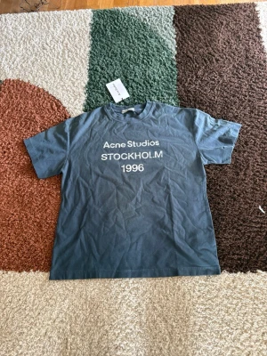 Blå Acne Studios t-shirt Stockholm - Snygg blå t-shirt från Acne Studios med tryck 'STOCKHOLM 1996' på bröstet. Klassisk passform och rund halsringning. Perfekt för dig som gillar stilrena och trendiga plagg med streetkänsla.