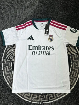 Real Madrid matchtröja Adidas vit - Säljer en vit Real Madrid matchtröja från Adidas med gröna detaljer och rosa ränder på axlarna. Tröjan har klubbmärke och Adidas-logga på bröstet samt sponsortryck 'Emirates Fly Better' framtill. Tillverkad i lätt och ventilerande material.