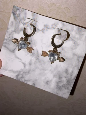Hjärtfomade örhängen med pil - Säljer ett par örhängen i guldtonad metall med hjärtformade stenar i silverfärg och pil-detaljer.