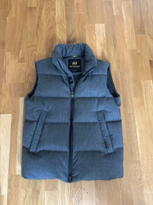 Hetrego väst   - Snygg grå dunväst från Hetrego med avtagbar huva och dragkedja framtill. Västen har två fickor med dragkedja och quiltad design som ger en puffig look. Perfekt för lager-på-lager och håller dig varm under kyliga dagar.
