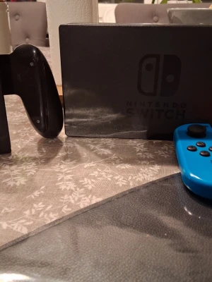 Nintendo Switch spelkonsol med blå Joy-Con - Nintendo Switch spelkonsol med svart dockningsstation och en blå Joy-Con-kontroll. Konsolen har en rektangulär form och skärmen är omgiven av svarta kanter. Perfekt för gaming både hemma och på språng.