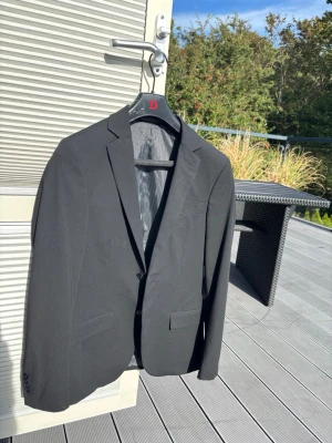 Svart kostym set  från Dressmann - Stilren svart kavaj från Dressmann med klassisk passform och två framfickor. Kavajen har smala slag och enkelknäppning. Fodret är randigt i grått och svart, vilket ger en snygg kontrast. Perfekt för dig som vill ha en clean och tidlös look. Byxorna ingår även med mått 30/30 straight/slim fit nypris 1400 galjan ingår.