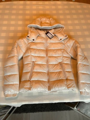 Vit Lager 157 jacka  - Snygg vit dunjacka med avtagbar huva och dragkedja framtill. Jackan har två snedställda fickor med dragkedja och quiltad design. Perfekt för kalla vinterdagar och ger en stilren look.