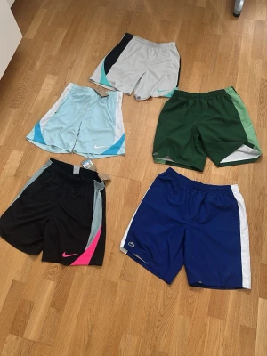 5-pack sportiga shorts Nike & Lacoste ”äkta” - Säljer ett 5-pack sportiga shorts från Nike och Lacoste i olika färger: svart med rosa detalj, blå med vit sida, grön med ljusgrön, ljusblå med vit och blå samt grå med svart. Perfekta för träning eller chill. Alla har elastisk midja och ledig passform. Värdet runt 2500kr