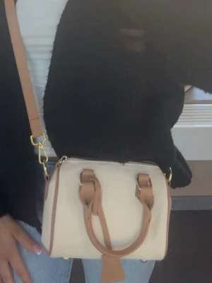 Vit och beige minihandväska med axelrem - Snygg minihandväska i vitt med beige detaljer och guldfärgade metalldelar. Väskan har både handtag och justerbar axelrem, cylinderformad design och dragkedja upptill. Nypris 165kr