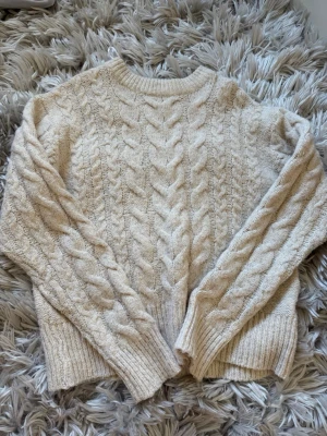 Beige kabelstickad tröja - Mysig beige kabelstickad tröja med rund hals och långa ärmar. Tröjan har breda flätmönster framtill och på ärmarna samt ribbade muddar vid ärmslut och nederkant. Perfekt för kalla dagar och enkel att matcha med jeans eller kjol.