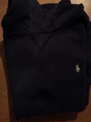 Mörkblå hoodie från Polo Ralph Lauren - Snygg mörkblå hoodie från Polo Ralph Lauren med klassisk huva och den ikoniska broderade loggan på bröstet. Hoodien har en avslappnad passform och är tillverkad i mjukt material som känns skönt mot huden. Perfekt för en chill och stilren look.