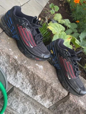 Nike TN - Säljer ett par Nike Tn sneakers i svart med röda och blå detaljer. Skorna har en sportig design med synliga Air Max-dämpningar i sulan, svarta snören och små vita swoosh-loggor på sidan. Materialet är en mix av textil och syntet.