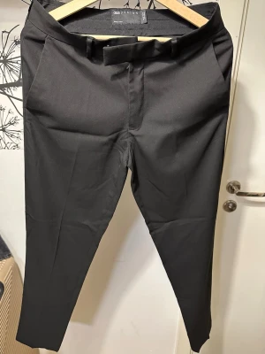 Svarta kostymbyxor från ASOS DESIGN - Helt nya svarta kostymbyxor från ASOS DESIGN med avsmalnande ben och hög midja. Byxorna har bälteshällor, sidofickor och en enkel dragkedja framtill. Perfekta för en clean och stilren look.
