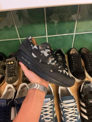 Nike Air Max 1 svart camo sneaker - Säljer ett par Nike Air Max 1 sneakers i svart med grått och vitt camouflagemönster. Skorna har klassisk snörning och en chunky sula med synlig Air-enhet. Materialet är en mix av mocka och textil vilket ger en schysst kontrast.