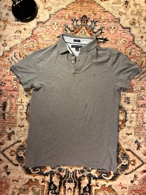 Grå pikétröja från Tommy Hilfiger - Säljer en klassisk grå pikétröja från Tommy Hilfiger med korta ärmar och liten logga på bröstet. Tröjan har krage och knappar framtill, perfekt till en avslappnad men ändå stilren look.