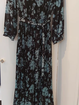 Lång blommig klänning med bälte - Säljer en navy klänning med ljusblått blommigt mönster. Klänningen har trekvartsärm, v-ringning och ett matchande bälte i midjan. Materialet är plisserat och ger en snygg struktur. Perfekt för dig som gillar en romantisk och trendig stil.