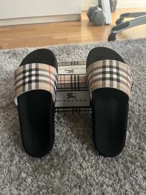 Burberry sandaler med rutigt mönster - Snygga sandaler från Burberry med klassiskt beige, svart och rött rutmönster på remmen och svart sula. Perfekta för sommaren och ger en lyxig touch till din outfit. Tillverkade i syntetmaterial med bekväm passform.